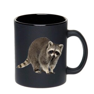 May include: A black ceramic coffee mug with a raccoon image printed on it. The raccoon is facing the camera and has a gray and black coat.