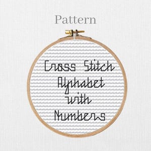 Op de afbeelding: Een kruissteekpatroon weergegeven in een houten borduurring. De tekst "Cross Stitch Alphabet with Numbers" is geborduurd met zwarte draad op een witte stoffen achtergrond. Het woord "Pattern" staat boven de ring.