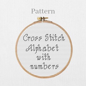 May include: A cross-stitch pattern in a wooden embroidery hoop. The text "Cross Stitch Alphabet with numbers" is stitched in black thread on a white fabric background. The word "Pattern" is above the hoop.