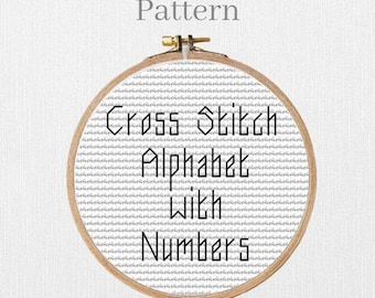 Puede incluir: Un patrón de punto de cruz en un bastidor de bordado de madera. El patrón presenta las palabras "Cross Stitch Alphabet With Numbers" en texto negro sobre un fondo blanco. La palabra "Pattern" está encima del bastidor.