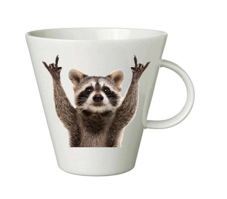 May include: White ceramic coffee mug with a raccoon wearing a mischievous expression and giving the peace sign with both hands. The raccoon is printed on the mug.