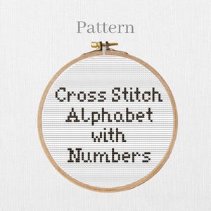 Op de afbeelding: Een kruissteekpatroon in een houten borduurring. De tekst "Cross Stitch Alphabet with Numbers" is met zwarte draad op witte stof gestikt. Het woord "Pattern" staat boven de ring.