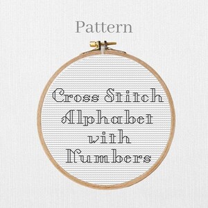 May include: A wooden embroidery hoop holding a cross-stitch pattern. The pattern features the text "Cross Stitch Alphabet with Numbers" in a decorative font. The word "Pattern" is above the hoop.