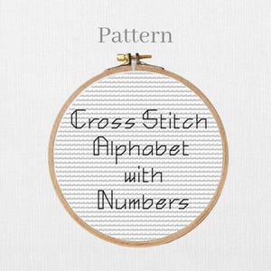 May include: A circular wooden embroidery hoop containing white Aida fabric with a grid pattern. The text "Pattern" appears above the hoop, and the text "Cross Stitch Alphabet with Numbers" is centred on the fabric in a black geometric font.