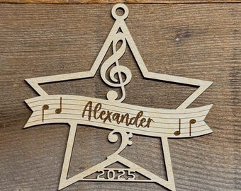 May include: A wooden star-shaped ornament with a musical theme. The ornament features a treble clef, musical notes, and the name "Alexander" on a banner. The year "2025" is also included.