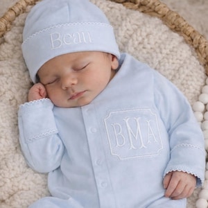 May include: A sleeping baby wearing a light blue footed onesie and matching cap, both embroidered with white lettering. The onesie features a monogrammed pocket. The baby is nestled in a woven basket on a soft, textured blanket.