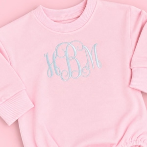 May include: A light pink long-sleeve romper featuring a ribbed crew neckline and ribbed cuffs. The center of the chest displays an embroidered monogram with the letters HBM in a light blue cursive script. The bottom includes three snap closures.