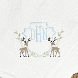 Peut inclure: Une couverture matelass&eacute;e blanche avec une broderie repr&eacute;sentant deux cerfs se faisant face. Entre les cerfs se trouve un contour de bouclier bleu clair contenant les lettres monogramm&eacute;es DHN. Le motif est agr&eacute;ment&eacute; de branches feuillues vert clair.