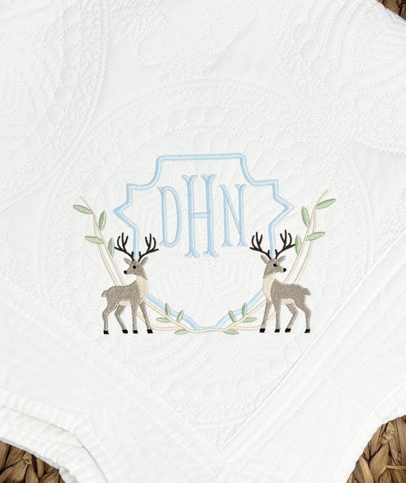 Peut inclure: Une couverture matelass&eacute;e blanche avec une broderie repr&eacute;sentant deux cerfs se faisant face. Entre les cerfs se trouve un contour de bouclier bleu clair contenant les lettres monogramm&eacute;es DHN. Le motif est agr&eacute;ment&eacute; de branches feuillues vert clair.
