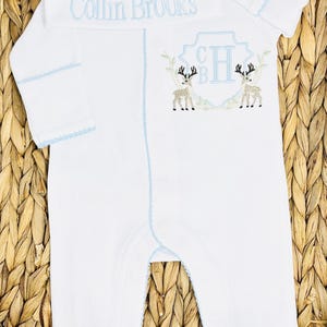 Peut inclure: Un ensemble pyjama blanc &agrave; manches longues avec pieds et bonnet assorti, dot&eacute; d'un liser&eacute; bleu clair. Le bonnet porte le nom brod&eacute; Collin Brooks. Le pyjama pr&eacute;sente un &eacute;cusson brod&eacute; avec les initiales CH et deux cerfs sur la poitrine.