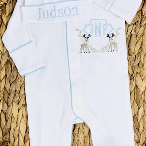 Peut inclure: Une grenouill&egrave;re blanche &agrave; manches longues avec pieds et un bonnet assorti &agrave; bordure bleu clair. Le bonnet porte le nom brod&eacute; Judson. La grenouill&egrave;re pr&eacute;sente un &eacute;cusson brod&eacute; avec les initiales JNP flanqu&eacute;es de deux cerfs marron et gris.