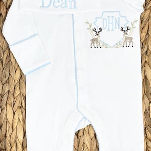 Peut inclure: Une grenouill&egrave;re blanche &agrave; manches longues avec pieds et un bonnet assorti, orn&eacute;s d'un liser&eacute; bleu clair. Le bonnet porte le pr&eacute;nom Dean brod&eacute; en bleu. La grenouill&egrave;re pr&eacute;sente un &eacute;cusson brod&eacute; avec les initiales DHN et deux cerfs.