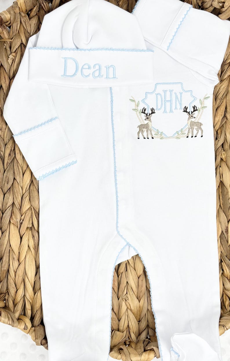 Peut inclure: Une grenouill&egrave;re blanche &agrave; manches longues avec pieds et un bonnet assorti, orn&eacute;s d'un liser&eacute; bleu clair. Le bonnet porte le pr&eacute;nom Dean brod&eacute; en bleu. La grenouill&egrave;re pr&eacute;sente un &eacute;cusson brod&eacute; avec les initiales DHN et deux cerfs.