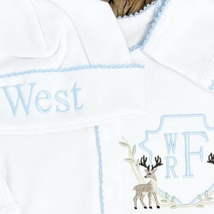Peut inclure: Un ensemble pour b&eacute;b&eacute; compos&eacute; d'un bonnet et d'un v&ecirc;tement &agrave; manches longues blancs avec des bordures bleu clair. Le bonnet porte le nom West brod&eacute; en bleu clair. Le v&ecirc;tement pr&eacute;sente un &eacute;cusson bleu clair avec les lettres WRF et deux cerfs.