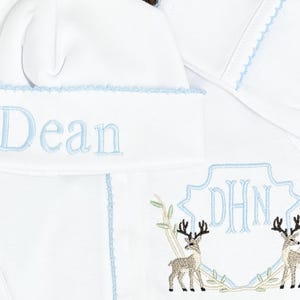 Peut inclure: Un bonnet de b&eacute;b&eacute; blanc et un v&ecirc;tement assorti avec des coutures d&eacute;coratives bleu clair. Le bonnet est brod&eacute; du pr&eacute;nom Dean en bleu clair. Le v&ecirc;tement pr&eacute;sente un monogramme en forme de bouclier bleu clair avec les lettres DHN et deux cerfs.