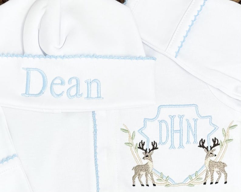 Peut inclure: Un bonnet de b&eacute;b&eacute; blanc et un v&ecirc;tement assorti avec des coutures d&eacute;coratives bleu clair. Le bonnet est brod&eacute; du pr&eacute;nom Dean en bleu clair. Le v&ecirc;tement pr&eacute;sente un monogramme en forme de bouclier bleu clair avec les lettres DHN et deux cerfs.