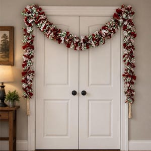 May include: A decorative garland draped over a double door frame, featuring a mix of fabric strips in red, white, and green. The garland includes patterns such as gingham and stripes, with long, light-colored fabric tassels hanging from each end.