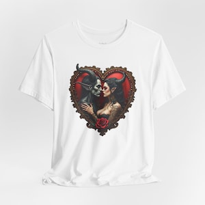 May include: White t-shirt featuring a heart-shaped graphic with a romantic gothic theme. The graphic depicts a dark-skinned couple with horns, embracing within a decorative frame, with a red rose at the bottom.