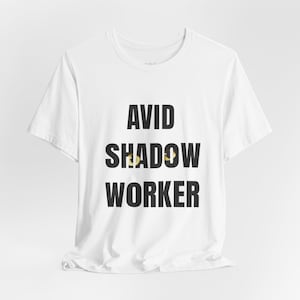 May include: White t-shirt with the words "AVID SHADOW WORKER" printed in bold black letters. The word "SHADOW" has a yellow and brown design in the middle. The shirt has a crew neck and short sleeves.