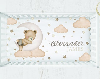 May include: A white cot sheet featuring a brown bear sleeping on a pink crescent moon. The sheet is decorated with beige clouds and gold stars. The text "Alexander James" is printed in brown on the sheet.