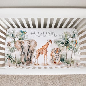 May include: A white crib with a fitted sheet featuring a jungle theme. The sheet has a watercolor design of an elephant, giraffe, tiger, palm trees, and parrots. The name "Hudson" is written in gray script on the sheet.