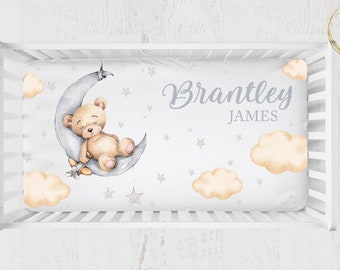 May include: A white cot sheet with a watercolour illustration of a brown teddy bear sleeping on a grey crescent moon. The sheet also features grey stars and yellow clouds. The text "Brantley James" is written in grey cursive lettering.