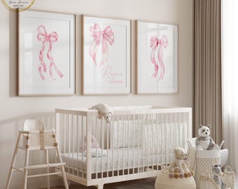 May include: Three framed watercolor prints with pink bow illustrations hang above a white crib. The middle print has the name "Regre Kincade" in cursive. A white high chair and decorative baskets with stuffed animals complete the nursery setting.