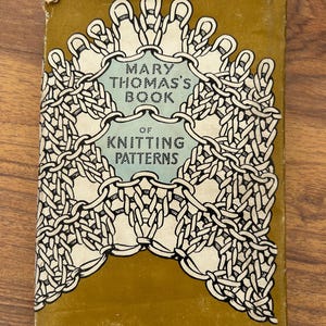Könnte beinhalten: Ein gebundenes Buch mit dem Titel Mary Thomas's Book of Knitting Patterns mit senfgelbem Einband. Die Vorderseite zeigt eine schwarz-weiße grafische Illustration von miteinander verbundenen Strickschleifen und Ketten um den zentrierten Text.