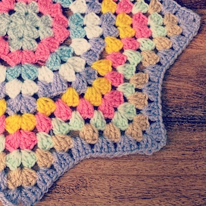 May include: A colorful crocheted granny square with a starburst pattern in shades of pink, yellow, blue, and green. The square is made with a variety of stitches and textures.