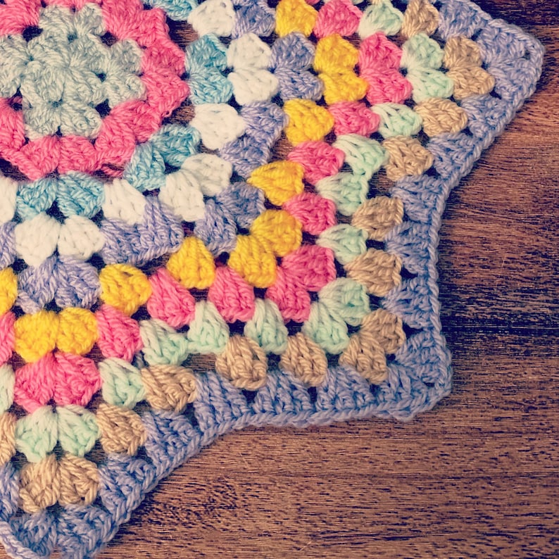 May include: A colorful crocheted granny square with a starburst pattern in shades of pink, yellow, blue, and green. The square is made with a variety of stitches and textures.