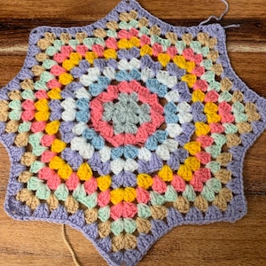 May include: A multi-colored crochet star with a center of grey, pink, blue, and white. The star is made with a variety of colors including purple, yellow, pink, green, and beige.
