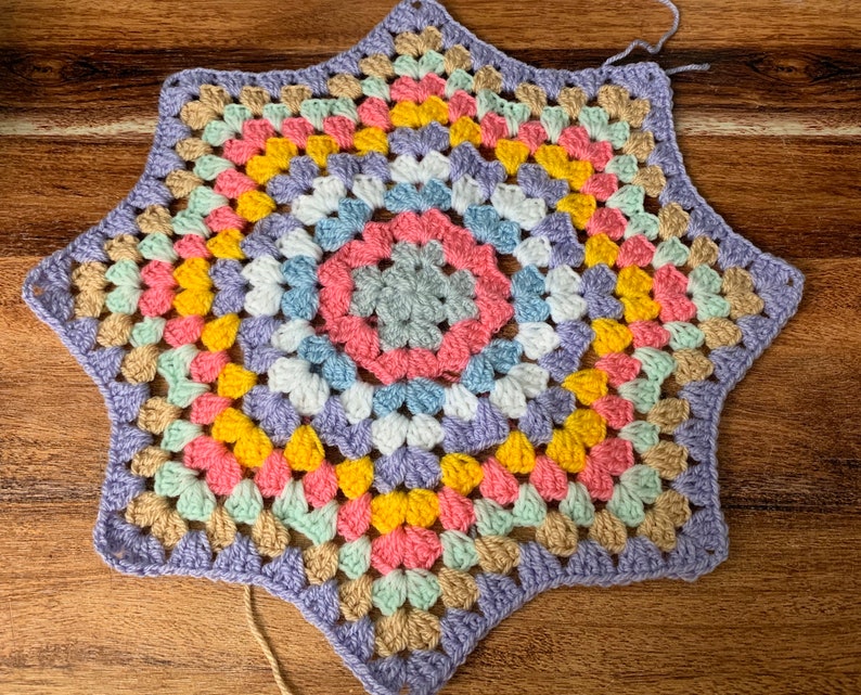 May include: A multi-colored crochet star with a center of grey, pink, blue, and white. The star is made with a variety of colors including purple, yellow, pink, green, and beige.