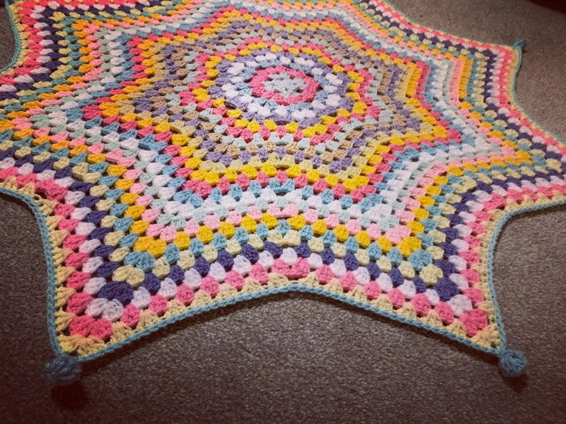 May include: A colorful crocheted star-shaped blanket with a multi-colored center and a light blue border.