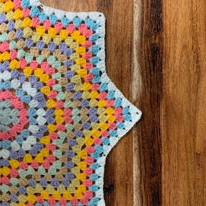 May include: A colorful crochet star-shaped coaster with a white border. The coaster is made with a granny square pattern and features shades of pink, yellow, blue, and green.