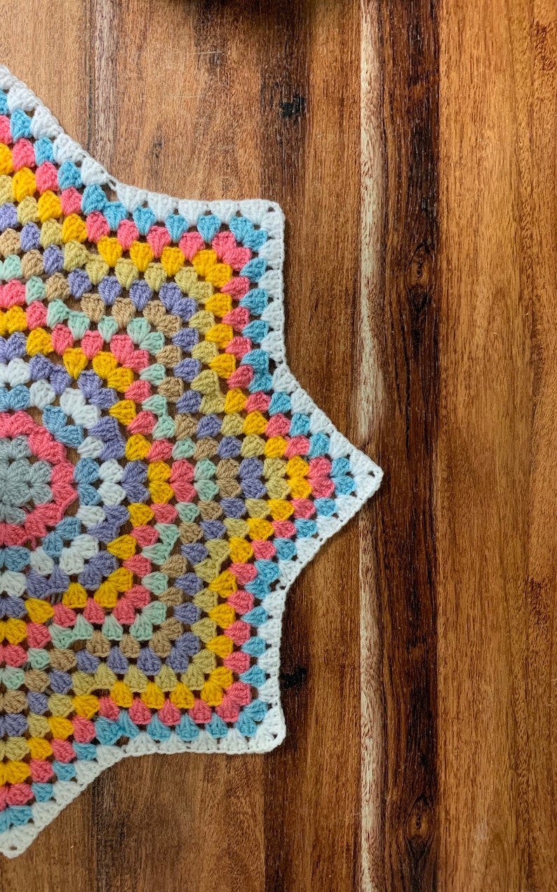May include: A colorful crochet star-shaped coaster with a white border. The coaster is made with a granny square pattern and features shades of pink, yellow, blue, and green.
