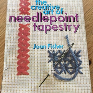 Puede incluir: Un libro titulado "The Creative Art of Needlepoint Tapestry" de Joan Fisher. La portada presenta un primer plano de puntadas de punto de aguja en hilo rojo y azul.