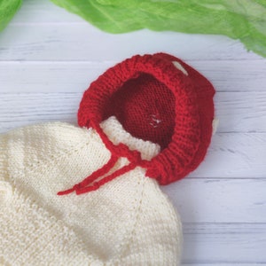 Peut inclure: Un pull en tricot blanc pour b&eacute;b&eacute; avec une capuche en tricot rouge. La capuche est surmont&eacute;e d'un pompon blanc.