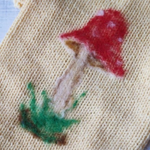 May include: A close-up of a knitted fabric with a felt mushroom design. The mushroom is red with white spots and has a brown stem. The stem is surrounded by green felt.