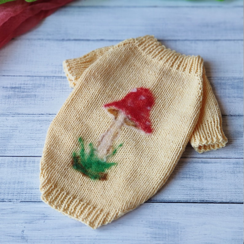 May include: A yellow knitted dog sweater with a red and white mushroom design. The mushroom has a green stem and green leaves.