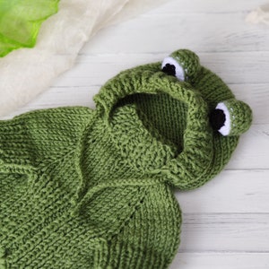May include: Green knitted baby frog hoodie with white eyes and a hood. The hoodie is made of soft yarn and is perfect for a newborn baby.