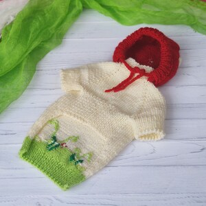 Peut inclure: Un pull en tricot de couleur cr&egrave;me avec une bordure verte et rouge et une capuche en tricot rouge. Le pull a un motif brod&eacute; vert et rouge sur le devant.
