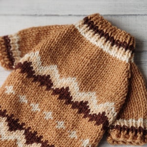 May include: A brown knitted jumper with a white and brown chevron pattern. The jumper has a ribbed collar and a loose fit.