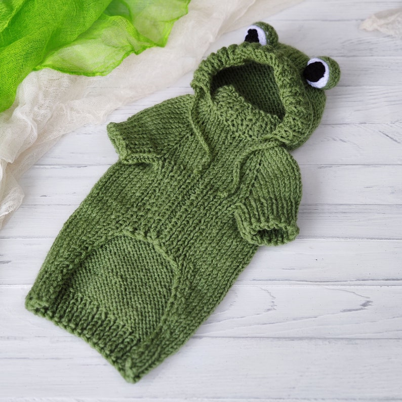 May include: Green knitted frog costume for a pet, featuring a hood with frog eyes and a  tail.