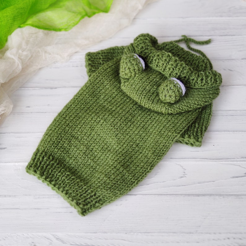 May include: Green knitted frog costume for a pet. The costume has a hood with a frog face and white eyes.