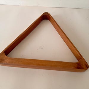 May include: A triangular billiard ball rack made of smooth, light brown wood with a natural grain pattern. The frame features three joined sides forming a hollow center, designed to hold and arrange billiard balls for a game of pool.