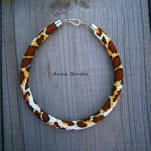 Puede incluir: Un collar de cuentas marrones, blancas y negras con un cierre plateado. El collar tiene forma de serpiente y está sobre una superficie de madera.