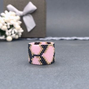 Peut inclure: Une large bague perlée avec un motif géométrique rose, noir et or. La bague est posée sur un fond gris, avec une boîte cadeau et des fleurs blanches en arrière-plan. Un bijou unique.
