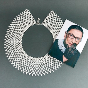 Pode incluir: Um colar de contas brancas no estilo de um colarinho, com um design delicado e intrincado. O colar está colocado sobre uma superfície cinza, com uma foto de Ruth Bader Ginsburg no fundo.