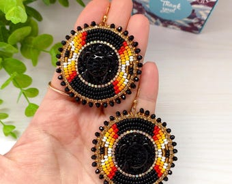 May include: Handmade beaded earrings with a black turtle design. The earrings feature a circular design with black, red, yellow, and white beads. The outer edge is lined with black beads and gold-colored accents.