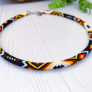 Puede incluir: Un collar circular de cuentas con un patrón geométrico repetitivo en cuentas de semillas negras, blancas, rojas y amarillas. El collar está construido con un diseño tubular tejido y cuenta con un cierre de metal plateado en la parte posterior.